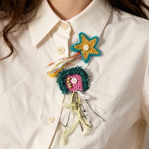 Op de afbeelding: Een set gehaakte broches bestaande uit een gele ster met een blauwgroene rand en een witte knoop in het midden, en een textuurrijke roze en donkergroene creatuurvorm met een witte knoop, versierd met bungelende veelkleurige garen- en lintstrengen.