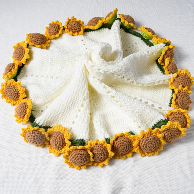 May include: A cream-colored, knitted baby blanket with a sunflower design. The blanket is bordered with crocheted sunflowers in yellow, brown, and green. The blanket is approximately 36 inches in diameter.