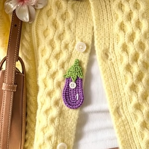 May include: A pale yellow cable-knit cardigan featuring a small, crocheted purple eggplant applique with a green stem and a white button detail. The cardigan is worn over a white top, and a brown leather bag strap is visible on the left side.