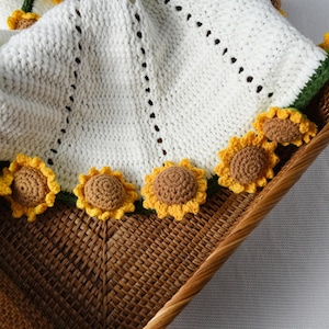 May include: A white crocheted blanket with a sunflower border. The blanket is made of a soft, textured yarn and features a scalloped edge. The sunflowers are yellow and brown, adding a cheerful touch to the design. The blanket is displayed in a wicker basket.