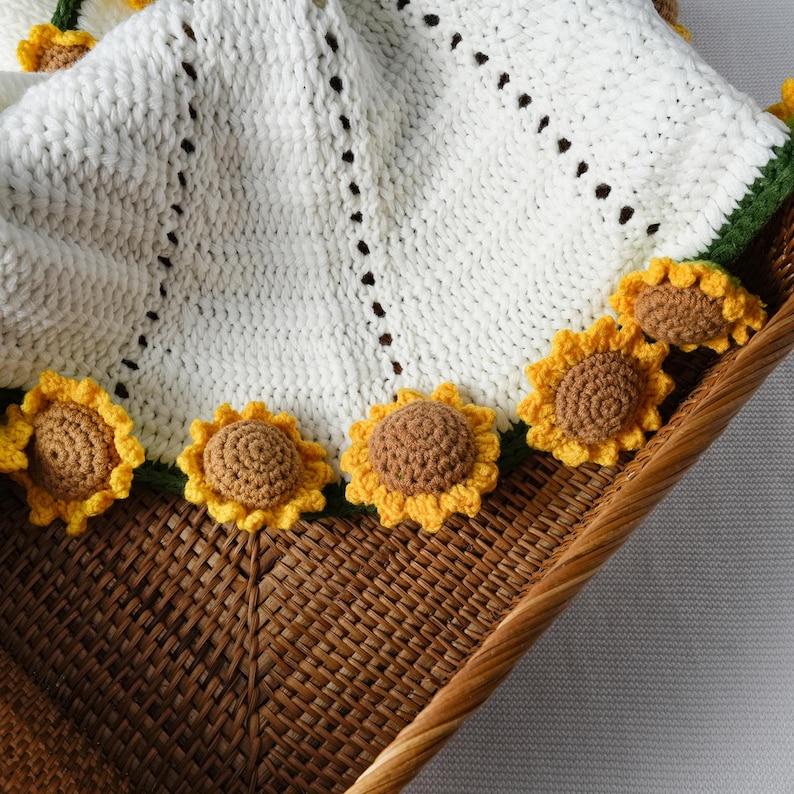 May include: A white crocheted blanket with a sunflower border. The blanket is made of a soft, textured yarn and features a scalloped edge. The sunflowers are yellow and brown, adding a cheerful touch to the design. The blanket is displayed in a wicker basket.