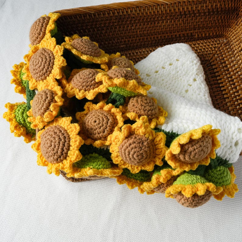 May include: A collection of handmade crochet sunflowers with yellow petals, brown centers, and green leaves. The sunflowers are arranged in a woven basket with a white crocheted item. The craftwork is detailed and vibrant.