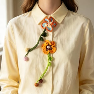 May include: A pale yellow button-down shirt featuring two crocheted flower appliques. The top flower has blue and orange petals with a white button centre. The bottom flower has orange petals with a brown centre and a white button.