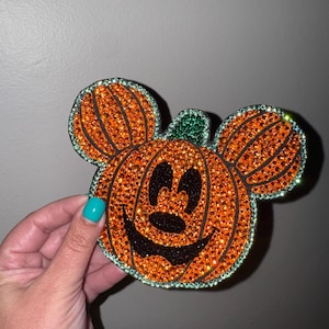 May include: A decorative patch shaped like a pumpkin with Mickey Mouse ears, entirely covered in sparkling orange, black, and green rhinestones. The design features a carved pumpkin face with black rhinestone eyes, nose, and a wide, grinning mouth.