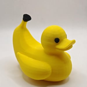 Puede incluir: Un objeto caprichoso en forma de pato amarillo con cuerpo de plátano. El pato tiene ojos negros y un pico amarillo. El cuerpo del plátano es curvo, con una punta negra. Este artículo único es una pieza decorativa lúdica.