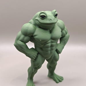 Puede incluir: Figura de rana verde y musculosa, con una cara sonriente y las manos en las caderas. La rana tiene músculos definidos y adopta una pose segura. La figura es un objeto sólido tridimensional.