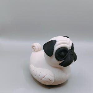 Puede incluir: Figura de un pug blanco con detalles negros en las orejas, ojos y nariz. El pug está sentado, con la cola enroscada y rasgos faciales detallados. La figura está hecha de un material liso.