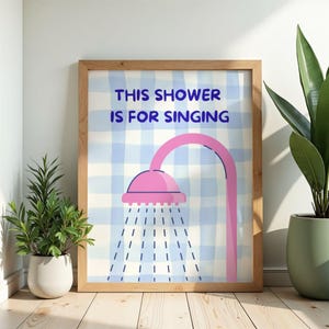 Könnte beinhalten: Ein gerahmter Druck mit dem Text "THIS SHOWER IS FOR SINGING" über einer rosa Duschkopf-Illustration. Der Hintergrund ist ein blau-weiß kariertes Muster. Der Rahmen ist hellbraun.