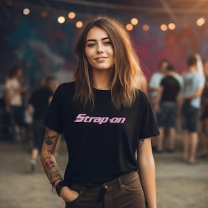 Peut inclure: Une femme portant un t-shirt noir avec le texte rose "Strap-on" imprimé sur le devant. Elle se tient devant un fond coloré avec des guirlandes lumineuses.
