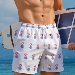 May include: White swim trunks featuring an all-over repeating pattern of pineapples with red and white striped tops and blue textured bases. The shorts have an elastic waistband with a white drawstring cord tied at the center front.