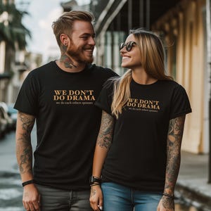 Peut inclure: Un couple portant des t-shirts noirs assortis avec le texte "WE DON'T DO DRAMA we do each others spouses" en orange. Le couple est debout dehors dans une rue de la ville.