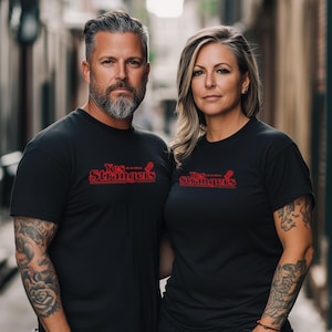 May include: Two people wearing black t-shirts with the red text "Yes...Strangers" and a microphone graphic. The man has a beard and tattoos on his arm. The woman has tattoos on her arm and blonde hair. They are standing in an alley.