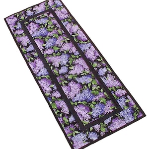 May include: A long rectangular runner with a black background and a floral pattern of purple and green lilac blossoms. The runner has a black border with a double black border.