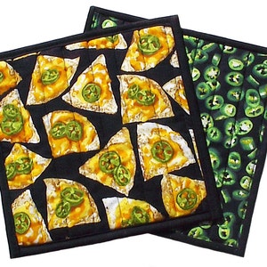 May include: Two black quilted pot holders with a black border. One pot holder has a pattern of yellow and green tortilla chips with jalapenos. The other pot holder has a pattern of green jalapeno slices.