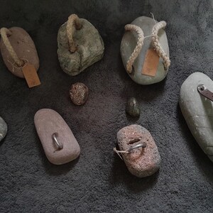 May include: A collection of nine unique stones, some with rope or leather accents, and some with metal rings. The stones are various shades of gray and brown.