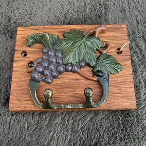 May include: A wooden wall-mounted key holder with a cast iron grapevine design. The grapevine features green leaves and purple grapes. The key holder has two hooks.