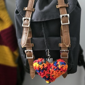 May include: A black backpack with brown faux leather straps and silver buckles, featuring a multi-colored crocheted heart keychain attached to the front. The heart is crafted from variegated yarn in shades of red, orange, yellow, blue, and purple.