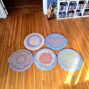 May include: Five handmade, circular rugs in various pastel colors, including blue, pink, and green, arranged on a wooden floor. Each rug features a spiral design, creating a textured appearance. The rugs are of varying sizes and are suitable for home decor.
