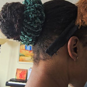May include: A close-up of a person's hair styled with a dark green, textured scrunchie. The hair is dark with a black headband. The person is wearing a black necklace.