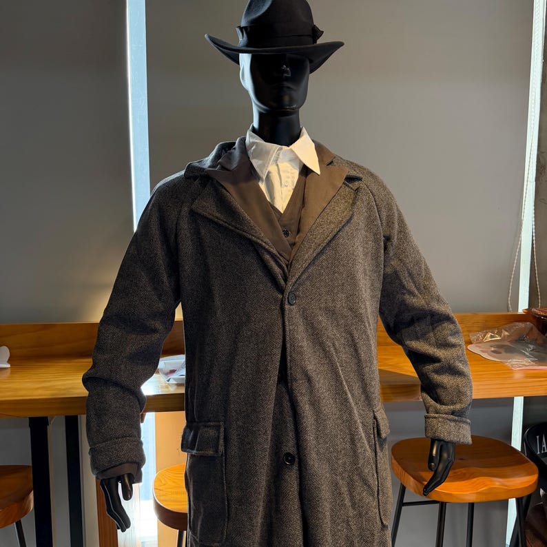 May include: A mannequin wearing a dark gray overcoat with a matching vest and a white collared shirt. The coat has large pockets and a single button closure. A black fedora hat completes the outfit. The mannequin is standing in front of a wooden table.