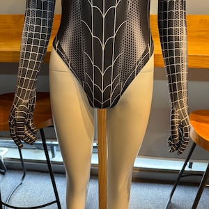 May include: Black and white bodysuit with a spiderweb design. The long sleeves and torso feature a web pattern, while the body has a stylized web design. The bodysuit is displayed on a mannequin.