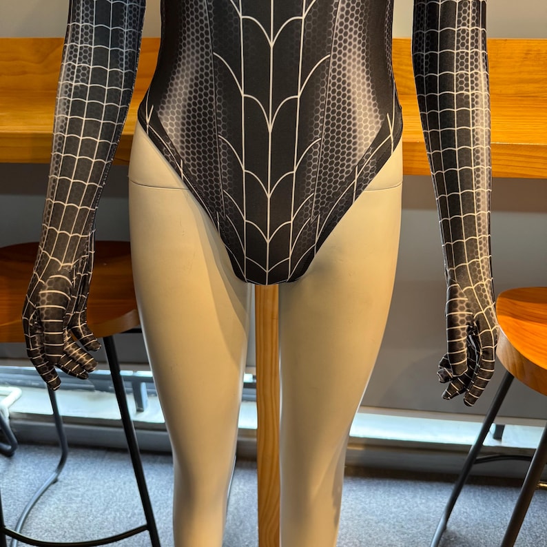 May include: Black and white bodysuit with a spiderweb design. The long sleeves and torso feature a web pattern, while the body has a stylized web design. The bodysuit is displayed on a mannequin.