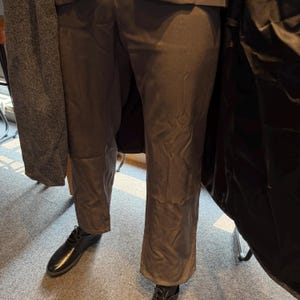 May include: A person wearing dark brown trousers and black leather shoes. A dark gray coat is draped over the person's arm. The trousers have a slight flare at the bottom.