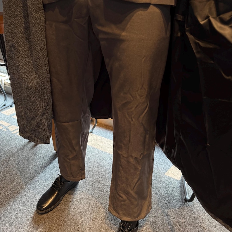 May include: A person wearing dark brown trousers and black leather shoes. A dark gray coat is draped over the person's arm. The trousers have a slight flare at the bottom.