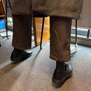 May include: Black leather dress shoes with dark brown soles. The shoes are laced and worn with dark brown trousers and black socks. The trousers are a solid color and have a slight sheen. The image is taken from a low angle.