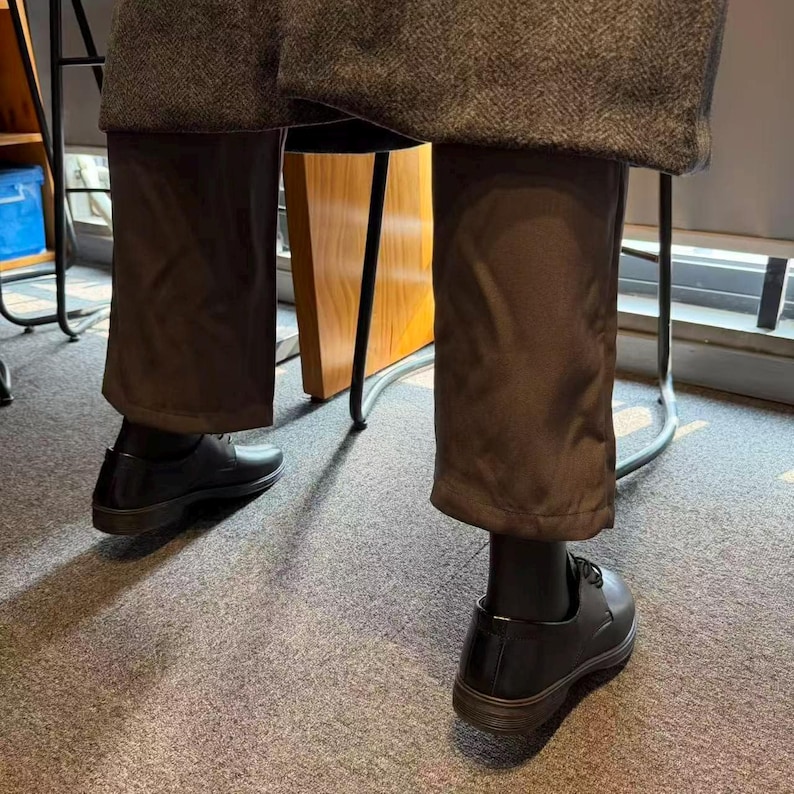 May include: Black leather dress shoes with dark brown soles. The shoes are laced and worn with dark brown trousers and black socks. The trousers are a solid color and have a slight sheen. The image is taken from a low angle.