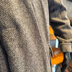 May include: Close-up of a gray herringbone pattern coat. The coat features a tailored design with a notched collar and a cuff on the sleeve. The fabric has a textured appearance.