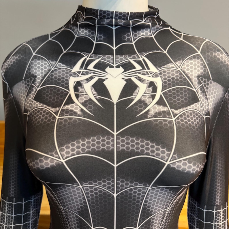 May include: A black and white bodysuit with a spiderweb pattern and a large white spider emblem on the chest. The long-sleeved garment has a high neckline and a detailed web design.