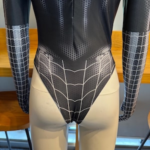 May include: Black bodysuit featuring a spiderweb design on the back and arms. The suit has a honeycomb pattern on the torso and arms. The arms are long-sleeved. The bodysuit is displayed on a mannequin.