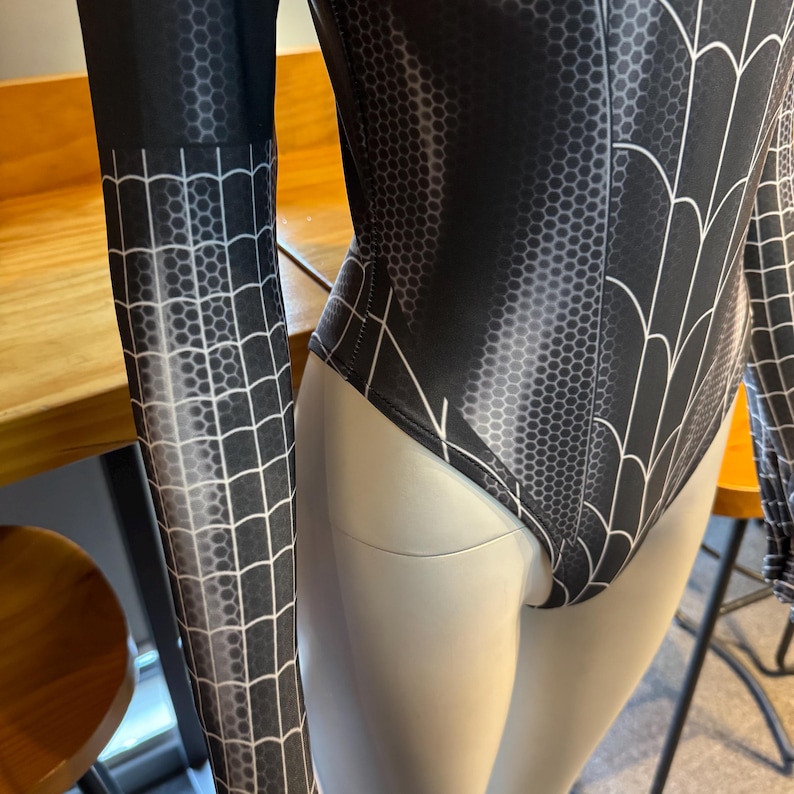May include: Black and white bodysuit with a spiderweb design. The long-sleeved garment features a honeycomb pattern and white web lines. The bodysuit is displayed on a mannequin.