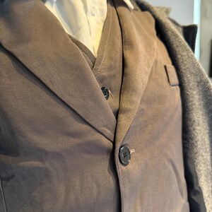 May include: A close-up of a brown suit jacket with a matching vest and a white collared shirt underneath. The jacket has two black buttons and a pocket. A gray overcoat is draped over the shoulders.