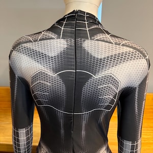 May include: Black and grey superhero costume with a spiderweb design. The back of the suit has a detailed honeycomb pattern and white accents. The suit features long sleeves and a zip down the back.