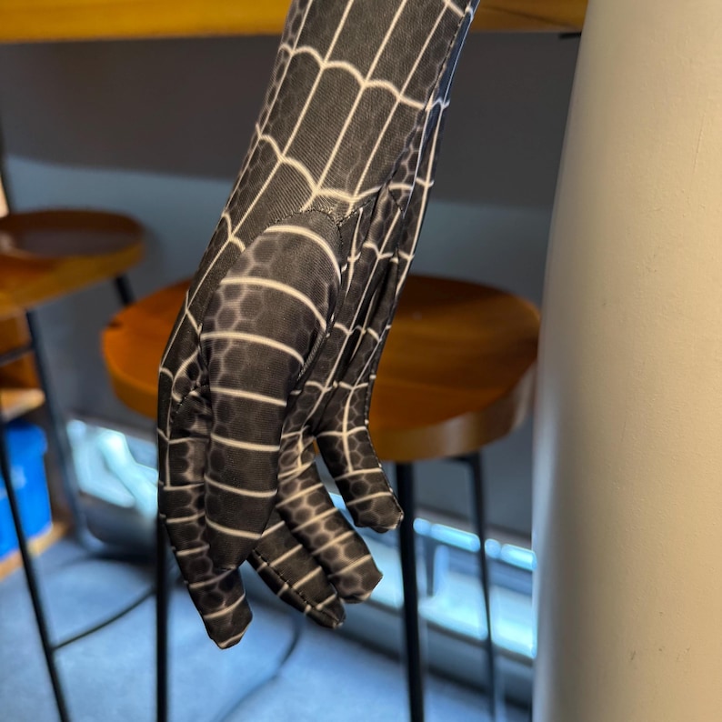 May include: Black and white patterned glove with a spiderweb design. The glove is made of a stretchy material and has a detailed web pattern. The fingers are visible, and the glove appears to be part of a costume.