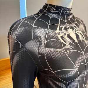 May include: Black and grey Spider-Man costume with a high collar. The costume features a web pattern and a white spider emblem on the chest. The fabric has a honeycomb texture.