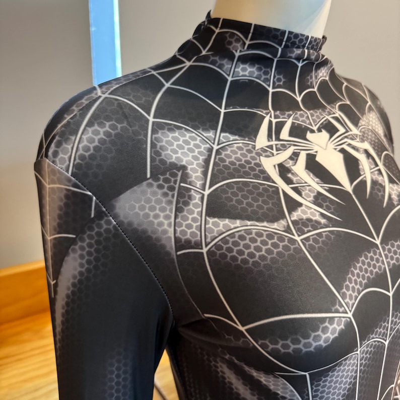 May include: Black and grey Spider-Man costume with a high collar. The costume features a web pattern and a white spider emblem on the chest. The fabric has a honeycomb texture.