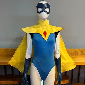 Può includere: Un costume da supereroe su un manichino. L'outfit presenta un body blu, un mantello giallo e una maschera abbinata. Il costume include anche lunghi guanti blu e un colletto giallo con una gemma rossa.