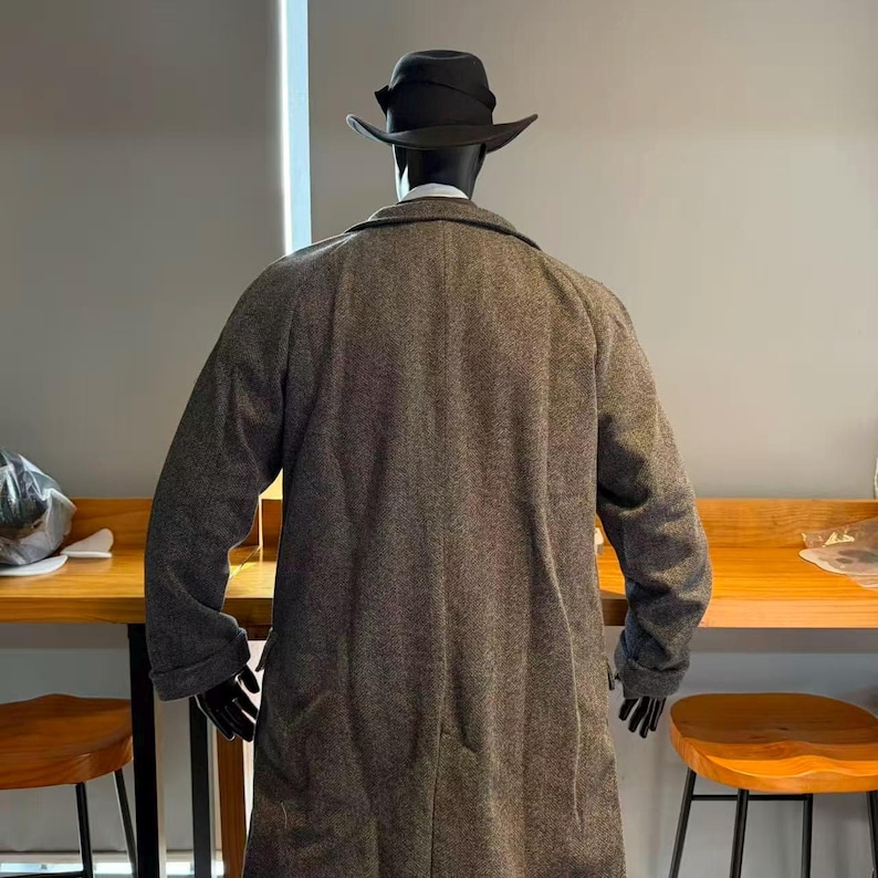 May include: A gray tweed overcoat with a black fedora hat on a mannequin. The coat is long, reaching below the knees, and has a classic, tailored design. The mannequin is positioned with its back to the viewer.