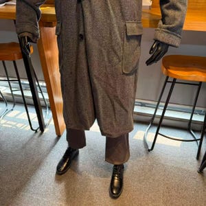 May include: A gray tweed overcoat with large pockets and a single button closure. The coat is paired with brown trousers and black leather shoes. The mannequin is wearing black gloves.