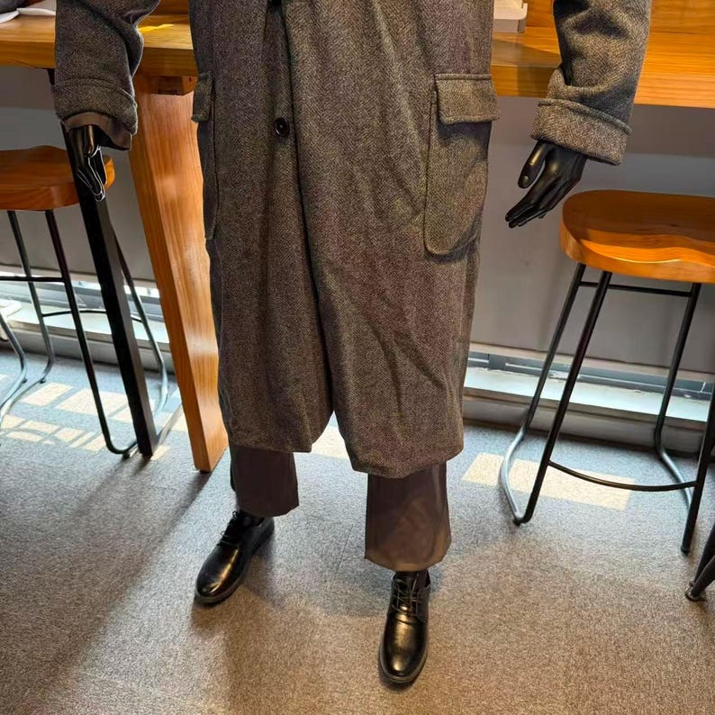 May include: A gray tweed overcoat with large pockets and a single button closure. The coat is paired with brown trousers and black leather shoes. The mannequin is wearing black gloves.