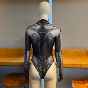 May include: A black and white bodysuit with a spiderweb design. The long-sleeved garment has a high neckline and a detailed pattern covering the entire suit. The design includes a web pattern on the front and back, with a gradient effect.