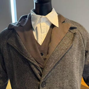 May include: A gray herringbone tweed coat with a brown lapel and a matching vest. A white collared shirt is visible underneath. The coat has a single button closure. The outfit is displayed on a mannequin.