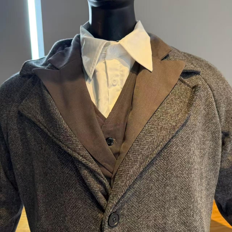 May include: A gray herringbone tweed coat with a brown lapel and a matching vest. A white collared shirt is visible underneath. The coat has a single button closure. The outfit is displayed on a mannequin.
