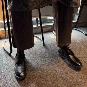 May include: Black leather dress shoes with laces. The shoes have a rounded toe and a low heel. The image shows the shoes from a low angle, with the legs of a person wearing brown pants visible above the shoes.