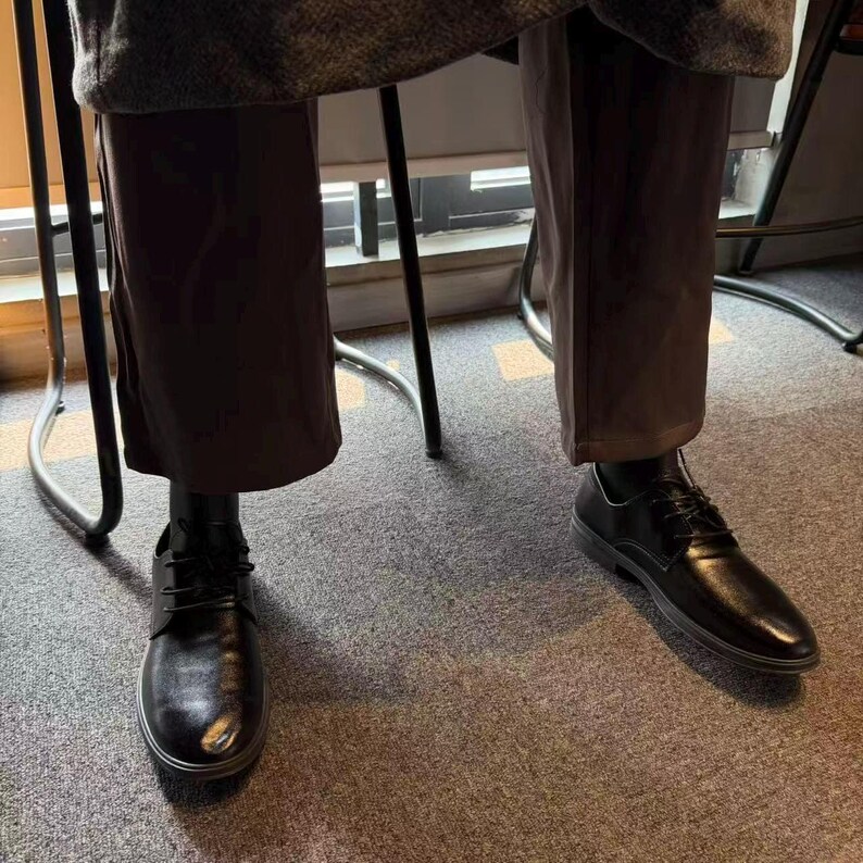 May include: Black leather dress shoes with laces. The shoes have a rounded toe and a low heel. The image shows the shoes from a low angle, with the legs of a person wearing brown pants visible above the shoes.