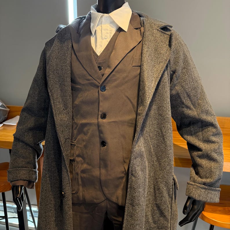 May include: A gray herringbone overcoat with a matching gray suit and a white collared shirt. The suit includes a vest and trousers. The coat is long and has a classic design. The mannequin is wearing black gloves.