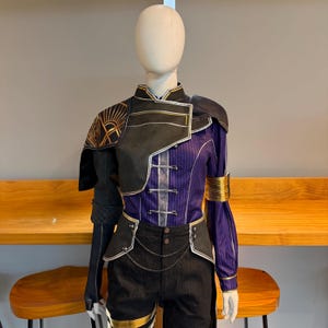 Puede incluir: Un disfraz exhibido en un maniquí. El atuendo presenta una parte superior morada y negra con detalles dorados, combinada con pantalones cortos negros. La parte superior tiene un cuello alto y hombreras decorativas. Los pantalones cortos tienen ribetes dorados y una cadena.